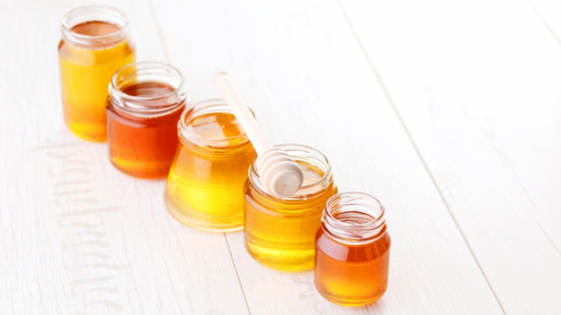 Pet tegli meda različitih nijansi poređanih u nizu na beloj podlozi, sa drvenom kašikom za med na jednoj tegli