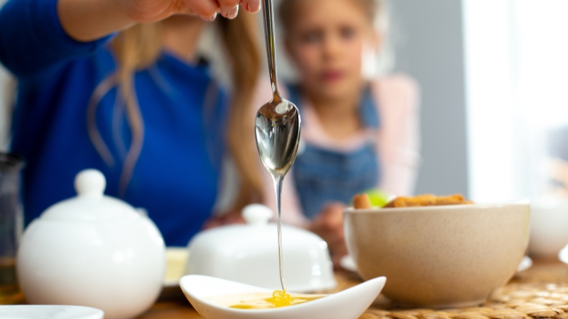 Kašika sa medom iznad male činije na stolu, dok dete u pozadini posmatra pripremu doručka