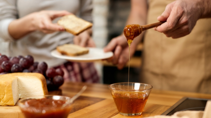 Osoba sipa med iz drvene kašike u staklenu činiju na kuhinjskom stolu, dok druga drži tanjir sa hlebom pored grožđa i sira