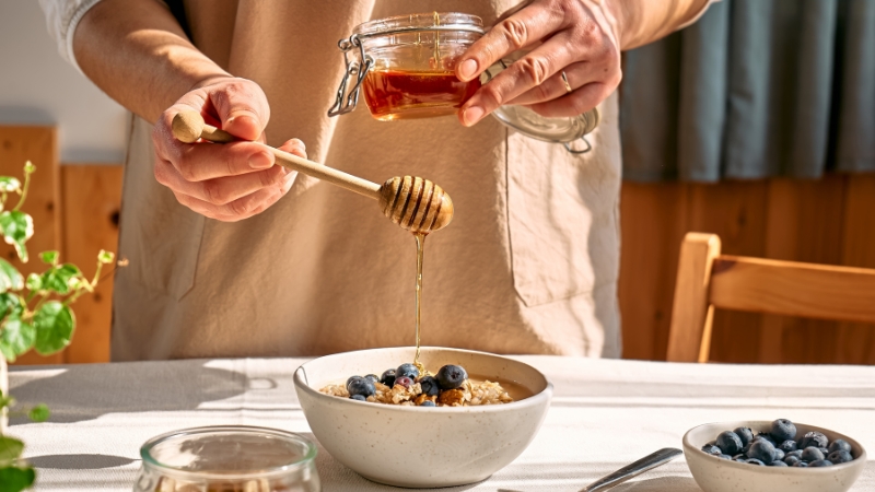 Osoba preliva med drvenom kašikom preko činije sa ovsenim pahuljicama i borovnicama